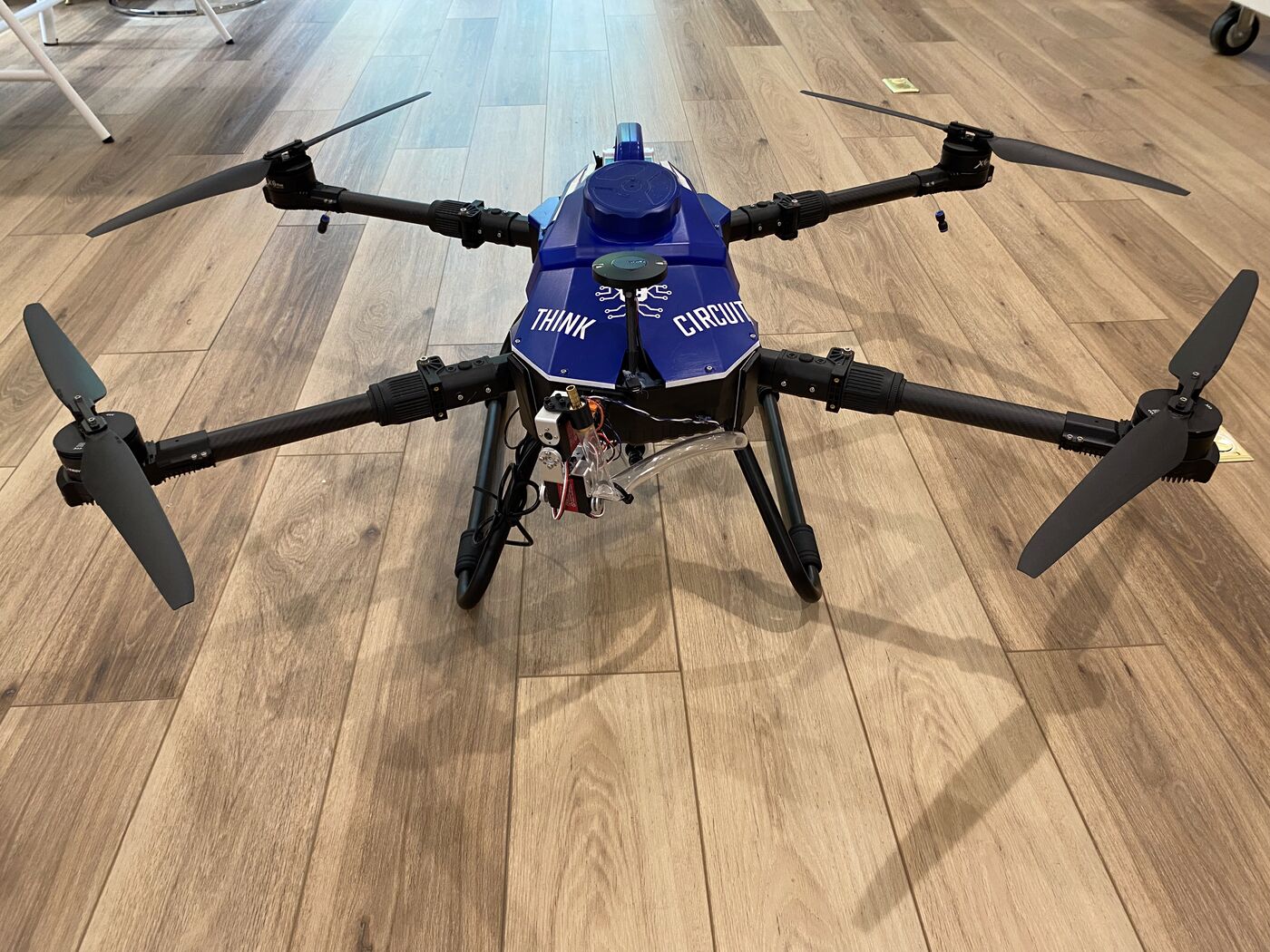 Three-quarter view of a blue Think Circuits suppression hexacopter on a wood floor, showing the slung water reservoir, pump, wiring, and a downward-facing sensor turret under the body.