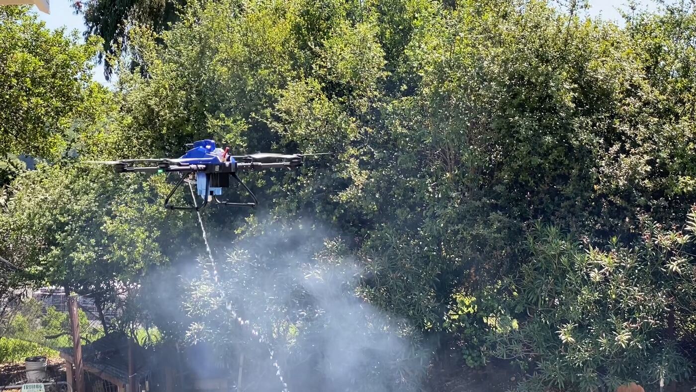 A black hexacopter drone hovering low over burning brush at the foot of a wood retaining wall, partly shrouded in steam, a stream of water arcing down from its nozzle into the flames.