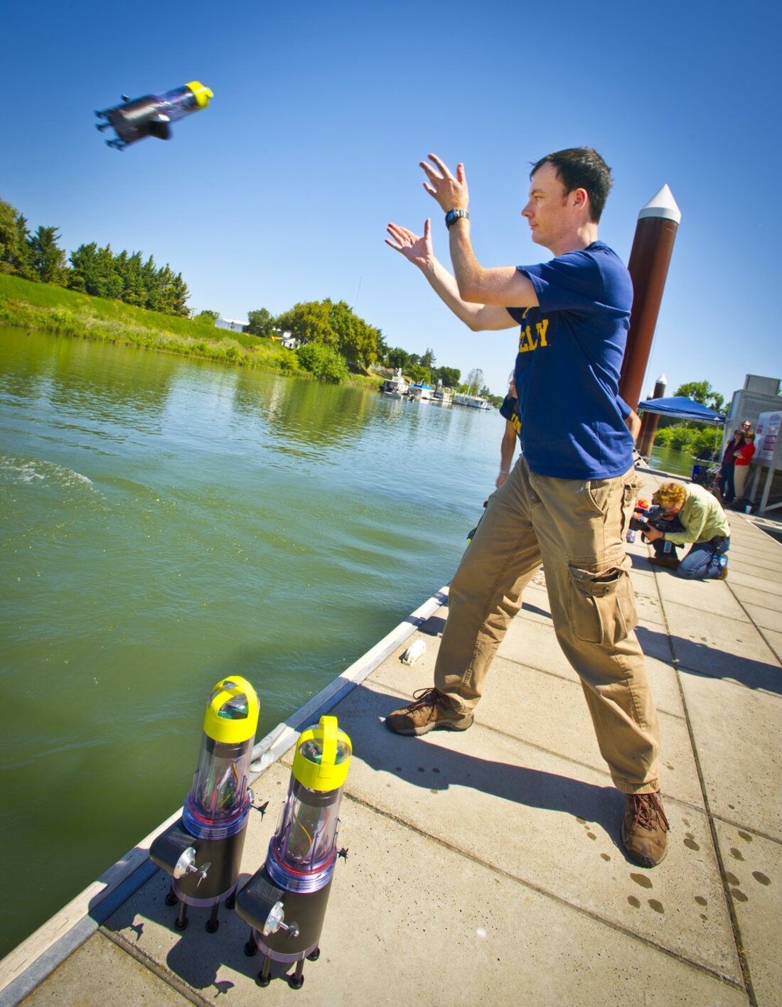 Researcher on a dock launching a self-propelled river drifter robot into the Sacramento–San Joaquin Delta, with low-cost Android-phone drifters in the foreground.